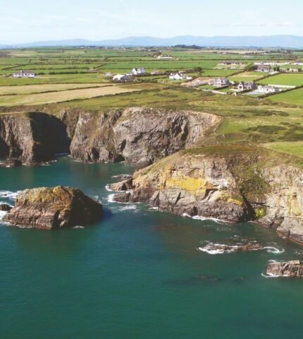 A picturesque coastal landscape in Waterford, Ireland, featuring rugged cliffs, calm blue sea, and green grass-covered land with sparse vegetation under a bright, clear sky.